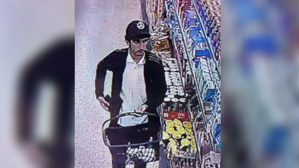 A CCTV image of a man in a black cap and jacket carrying a shopping basket
