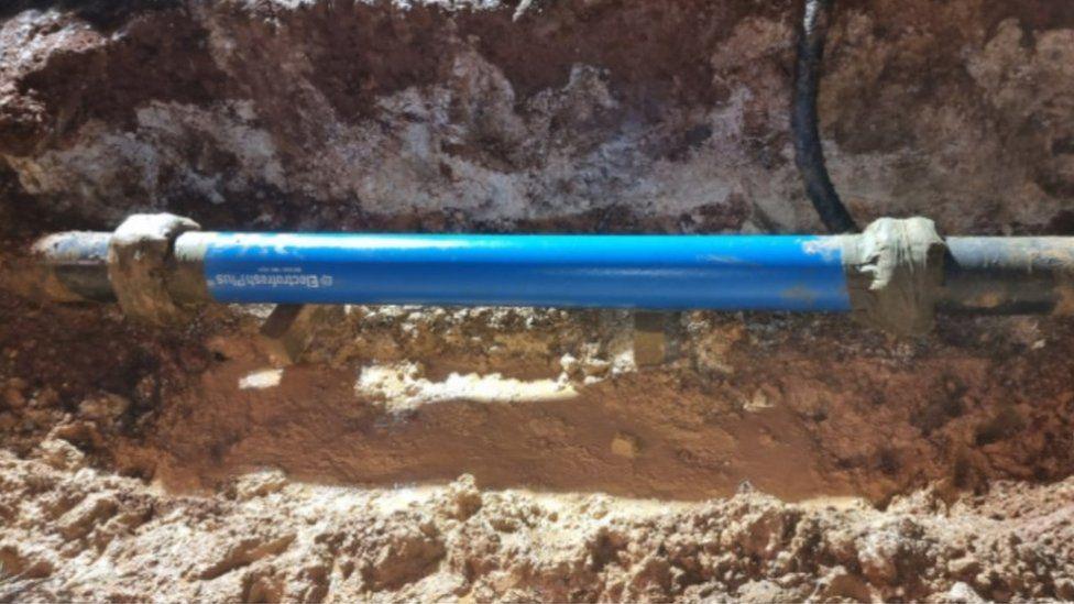 Hole in ground with sandy coloured earth and bright blue fixed water pipe in centre.