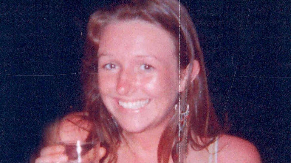Head and shoulders image of Lucy Hargreaves smiling at the camera and holding a drink against a black background