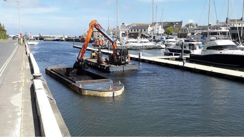 Peel Marina, there are modern boats along berths in the background,, in front you can see a vessel with a yellow crane scooping thick brown material into another vessel.