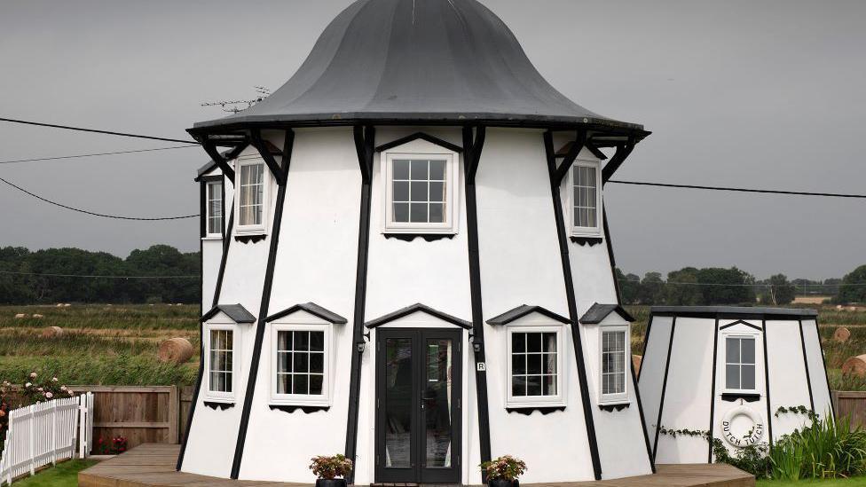 A converted helter-skelter. The main building is two storeys and has a black domed roof. The rest of the building is white with black vertical timber lines and a black door. On the side is another section of the old fairground ride. It is also black and white. 