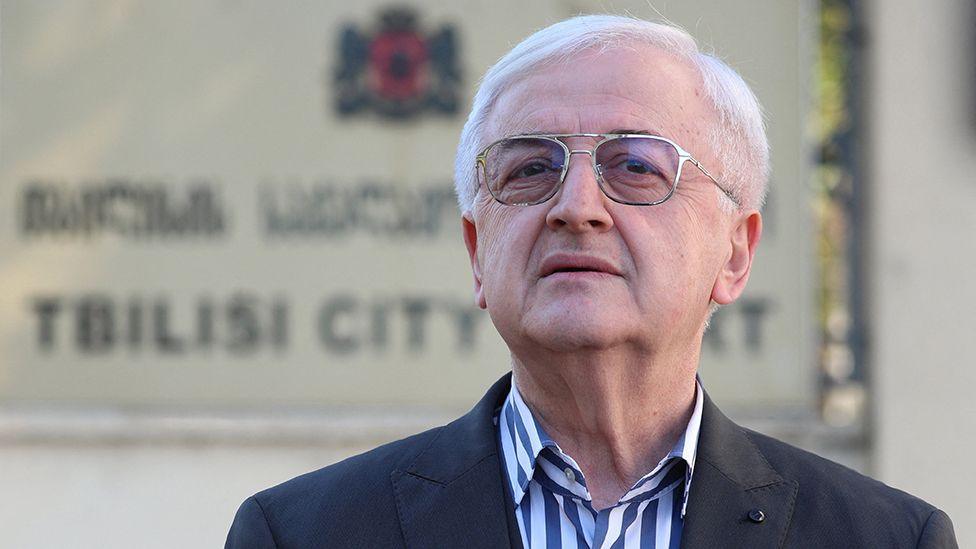 Miss Culley's lawyer, Malkhaz Salakaia, is standing in front of Tbilisi City Court in Georgia, though the sign on the wall behind him saying this is out of focus. He is wearing a black suit jacket and an open-necked shirt with thick stripes in dark blue and white. He has silver rimmed glasses and short, white hair and has his head held confidently upright, chin slightly jutting out.
