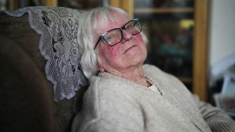 Jean, an elderly person with white hair is seated in a cushioned chair with a patterned fabric cover. The person is wearing a light beige knitted sweater. In the background, there is a wooden cabinet with glass doors displaying various items.