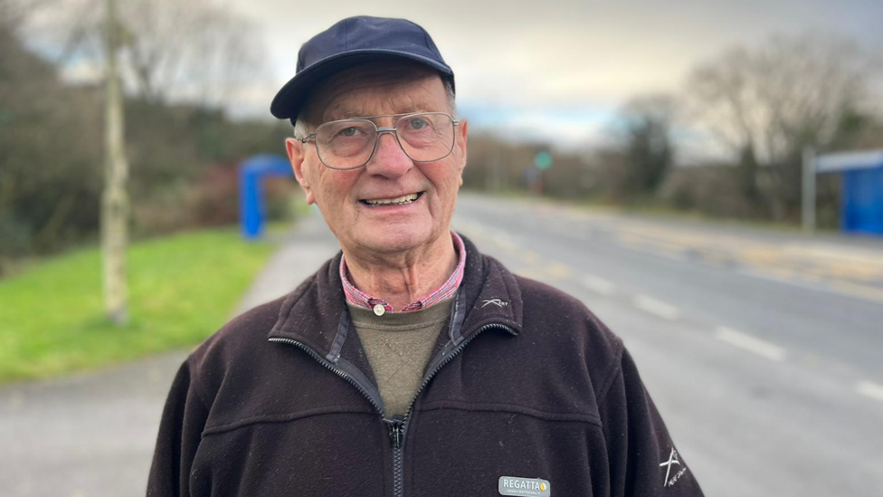 A man is smiling at the camera, he is wearing large silver-rimmed glasses. He has a dark blue hat on and is wearing a black zipped jumper, underneath he has a grey jumper and you can see the collars of a pink shirt peaking through. He is stood on the roadside and behind him is a blurry image of a grass verge to the left and road on the right. The man is the focus of the picture.