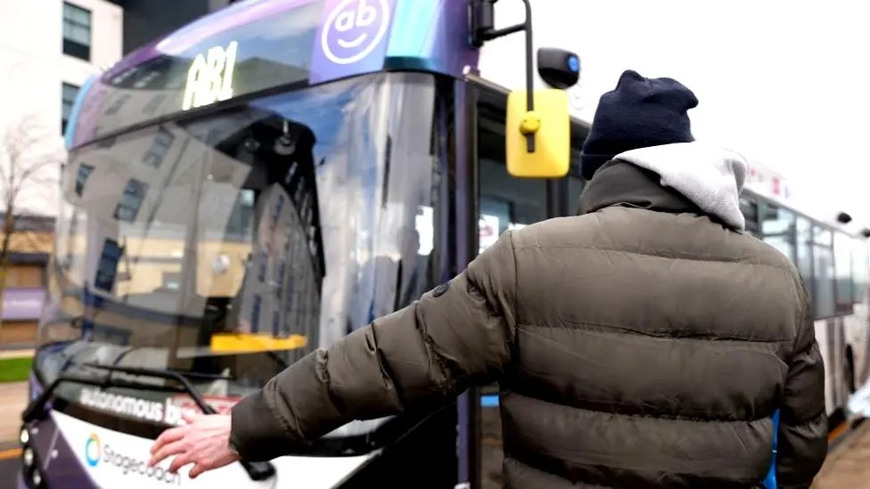 A man wearing a black padded jacket and a navy hat holds out his arm to hail an autonomous bus marked AB1. 