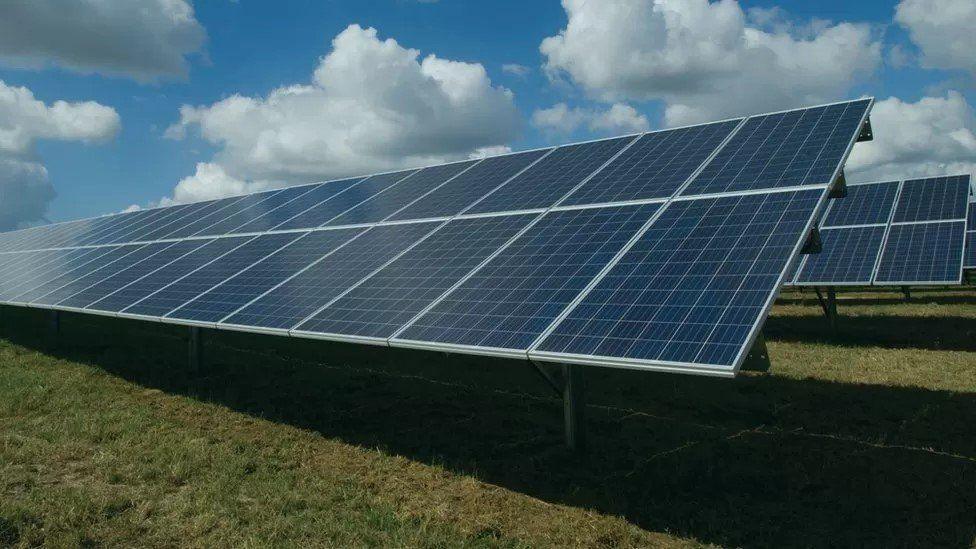 A view of large solar panels on legs set into close-cropped grass with a blue sky with white clouds above