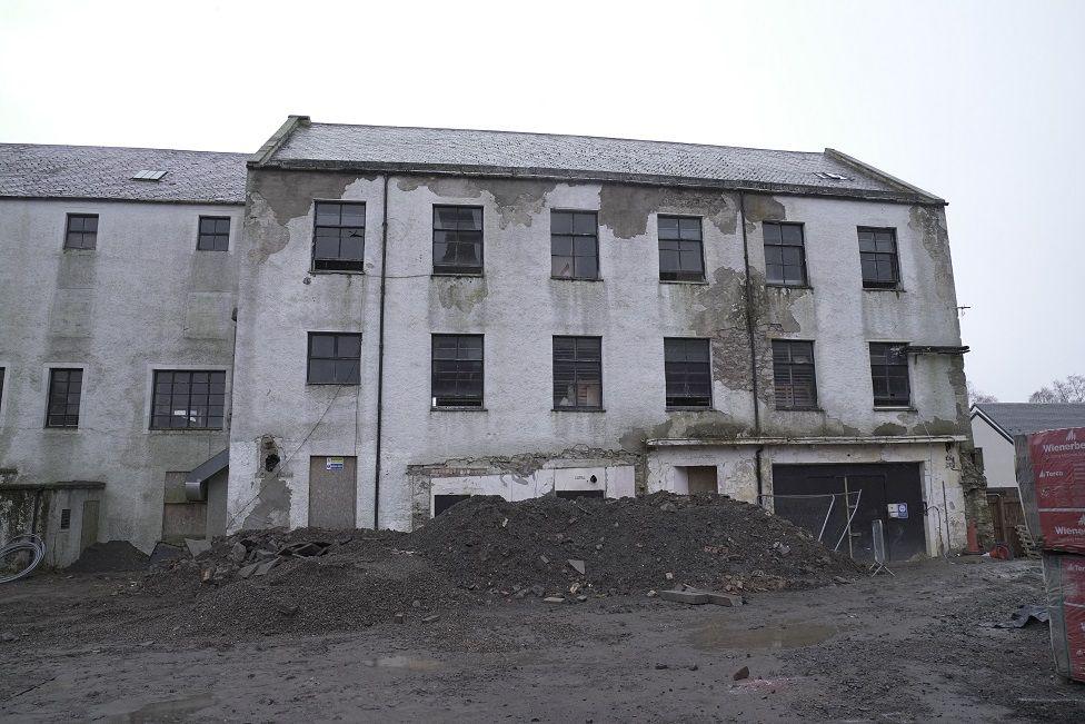 A dilapidated old mill building with plaster flaking off its walls and a pile of rubble outside it