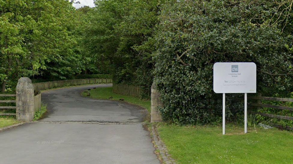Google Streetview image of the entrance to the school, showing a meandering asphalt drive bordered with a low stone wall and surrounded by large green trees. To the right before the entrance is a white sign for the school on a grassed area