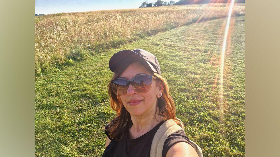 Geeta pictured on a gentle hill with green grass and then a field of taller grasses or crops. Rays of sun shine over the scene. She wears large brown sunglasses, a brown baseball cap, a black T-shirt and beige rucksack strap. Her hair is shoulder length and reddish/brown.