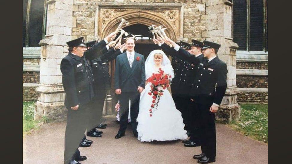 Mervyn Housden in a grey suit and red buttonhole standing beside Hayley Housden in a long white wedding dress and veil holding a bouquet of red flowers, outside a church with a guard of honour of firefighters on either side in dark uniforms and caps holding hammers aloft