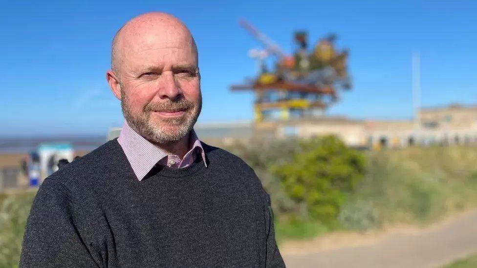 Mark Canniford looking at the camera with a beard and a dark jumper and shift on and a view of a beach in the distance, but out of focus