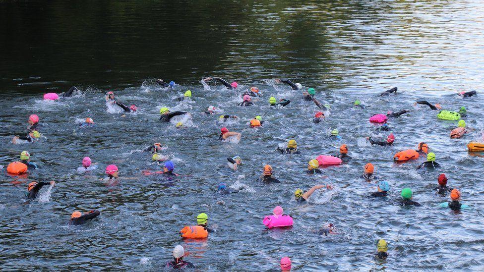 Dozens of swimmers with colourful swimming buoys and hants in orange, pink, yellow and blue - the water is choppy as the swimmers are racing along a course.