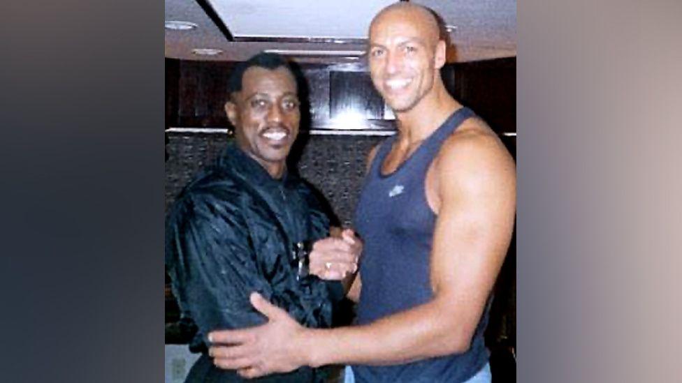 Daz Crawford posing for a photo with Wesley Snipes. Both are smiling and out of their movie costumes. Crawford, who is a few inches taller than Snipes, is wearing a blue vest. Snipes has a black jacket on.