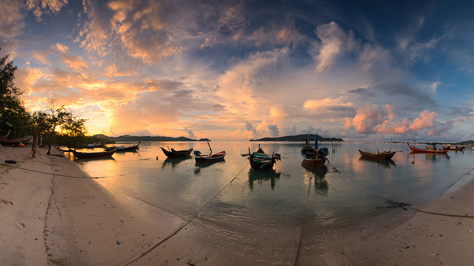Small boats are in the shallows of the sea off the coast of Chalong Bay in Phuket, with two pulled up on the shore. It is the early evening, with some mall islands in the distance