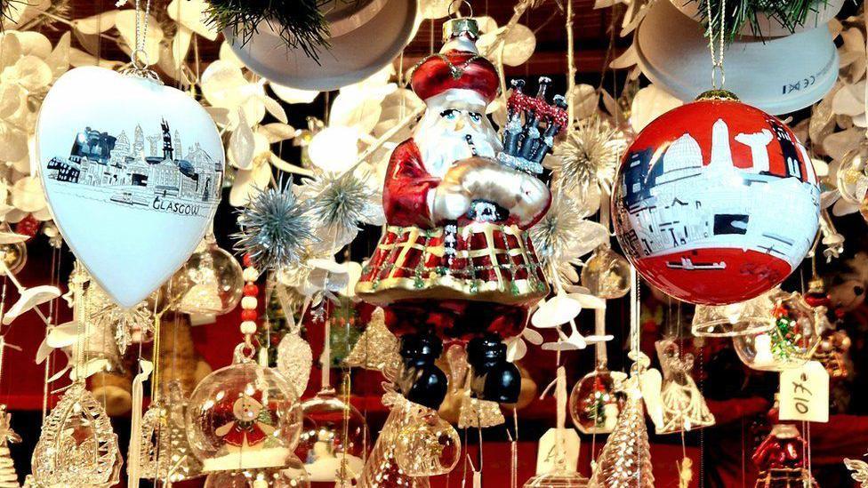 A close-up image of baubles hanging from a ceiling in a shop. You can see price tags hanging from some of them. There is a Santa holding bagpipes which is the most prominent bauble.