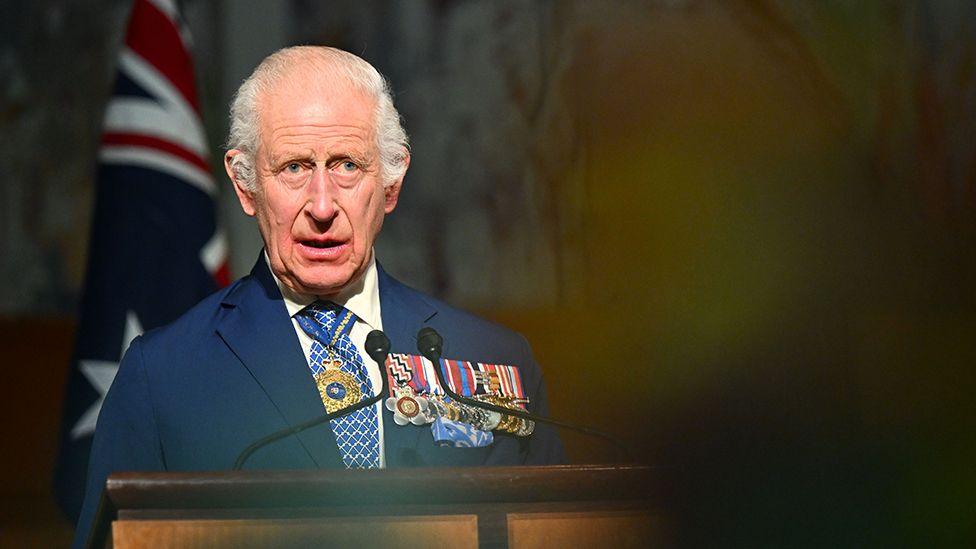 King Charles delivers a speech in the Australian Parliament on Monday,  he is wearing a suit and white shirt with a blue and white tie, with several medals on his left lapel and a gold necklace around his neck. He's looking slightly away from camera, with an out-of-focus audience member in the foreground and an Australian flag in the background.