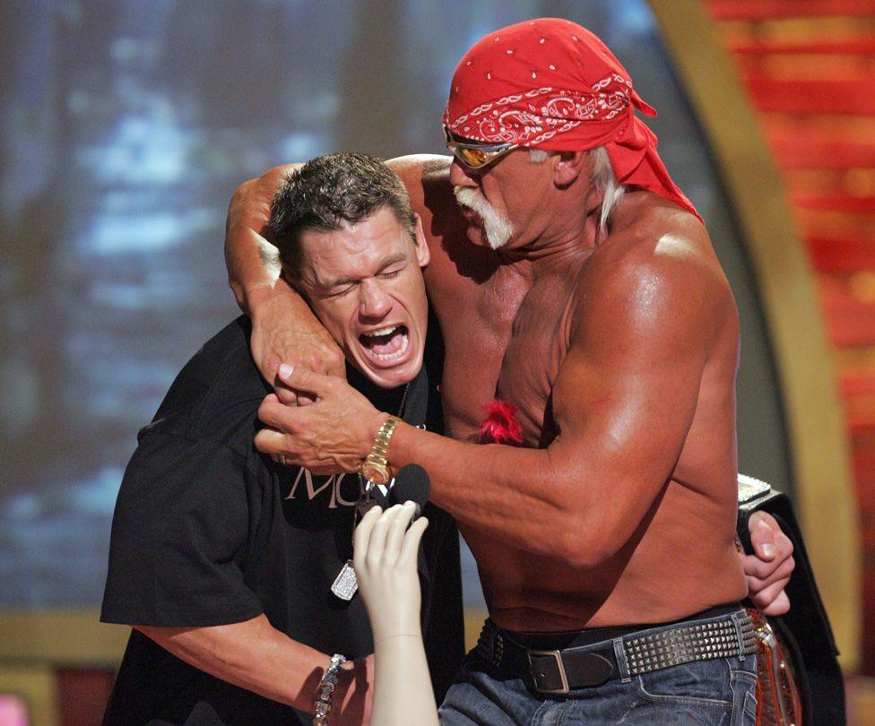 Hulk, bare chested and wearing a red bandana and sunglasses, with John Cena in a headlock during 2005 Teen Choice Awards in California. Cena is feigning a pained look.