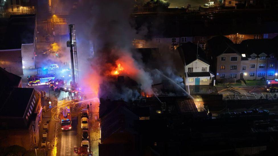 A drone shot of flames and large plumes of smoke coming from a building. There are large amounts of emergency service vehicles in the road next to the building. 