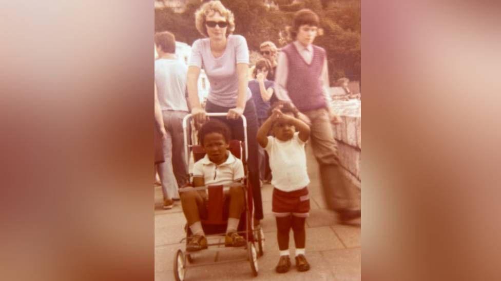 A photograph from the 1970s shows the family walking along a prom, with six year old Adrian in his younger brother's pushchair, while his three-year-old brother walks alongside.