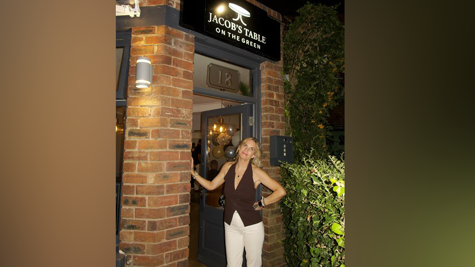A woman with blonde hair, wearing a brown halter-neck top and white trousers, leans in a doorway and smiles at the camera.