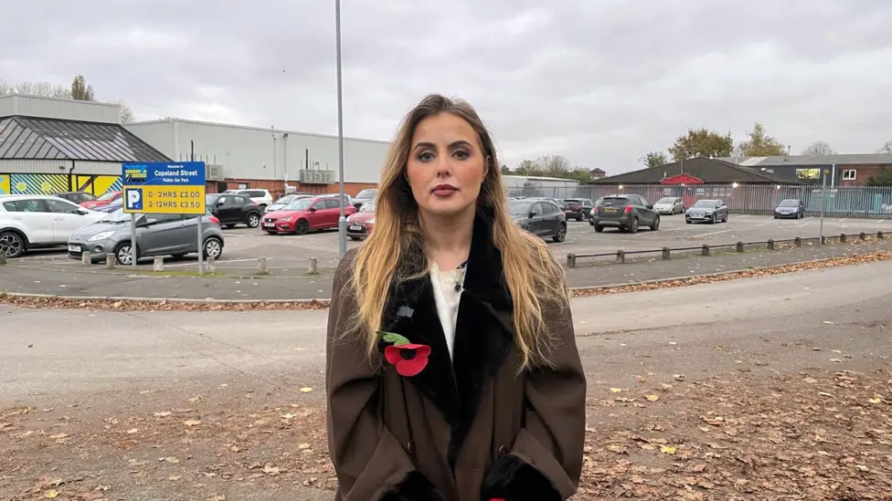 Miss Hudson, who is a young blonde woman, stands outside the Excel carpark in question, wearing a brown coat with a poppy on the lapel