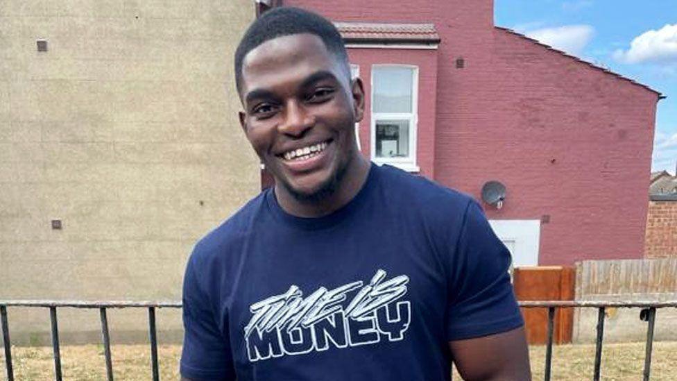 A medium close up shot of Chris Kaba outside, smiling and wearing a dark blue T-shirt.