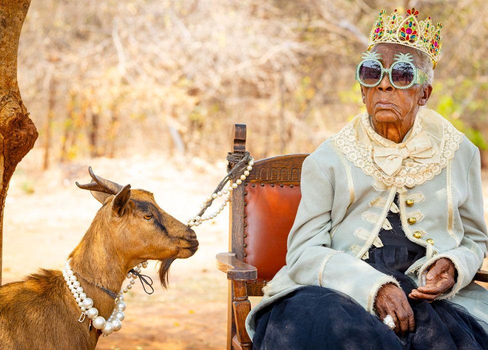 A goat wearing a big pearl necklace which tethers it to a wooden chair on which Margret Chola sits wearing a plastic bejewelled crown, sunglasses with bejewelled frames in the shape of pineapples and a creamy jacket with lace and big golden buttons over a dress made of black netting