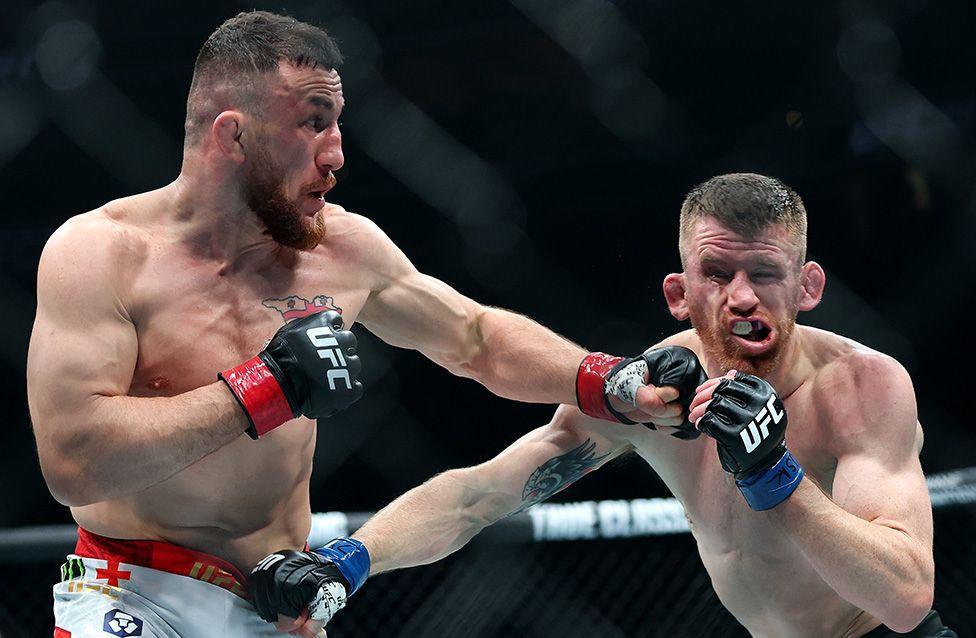 Merab Dvalishvili lands a punch against Cory Sandhagen during a bantamweight title bout at UFC 320 in Las Vegas. The image shows Dvalishvili in white shorts striking Sandhagen, who is wearing blue gloves, inside the octagon cage.