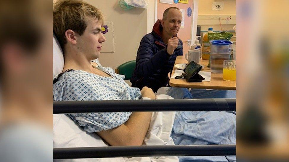 Jasper in a hospital bed, wearing a hospital gown with his father sitting next to him.