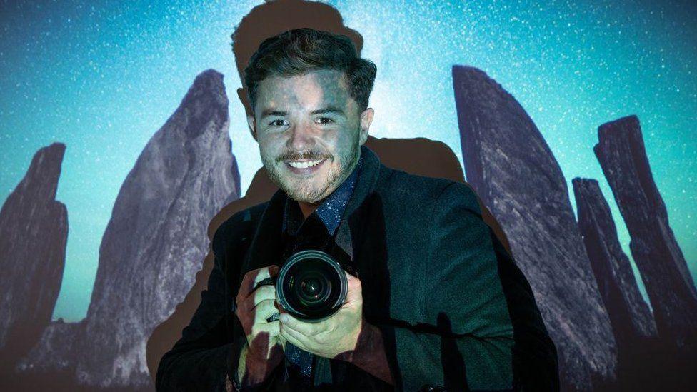 Josh Dury posing with his camera and smiling, a projector casts blue shadows over him. A picture of the Callanish stones is displayed over and around him.