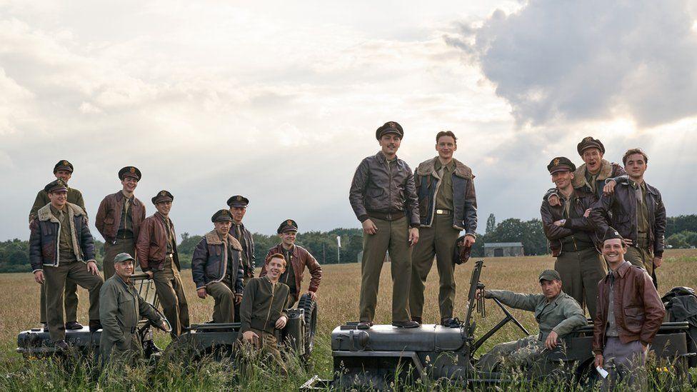 Masters of the Air publicity shot showing 16 airmen in World War Two uniforms and standing on military trucks in a field.