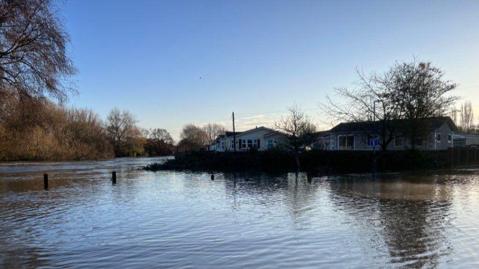 Wooden posts along river bank sumberged by water with tree lined river off to right and number of park homes alongside.