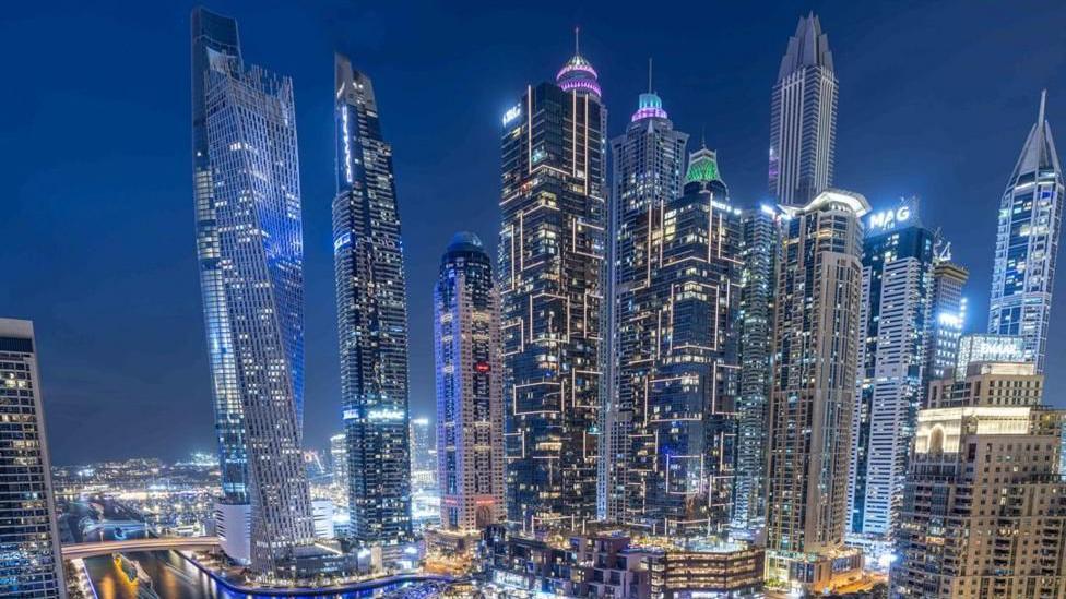 an aerial shot of Dubai at night, showing tall buildings and boats illuminated