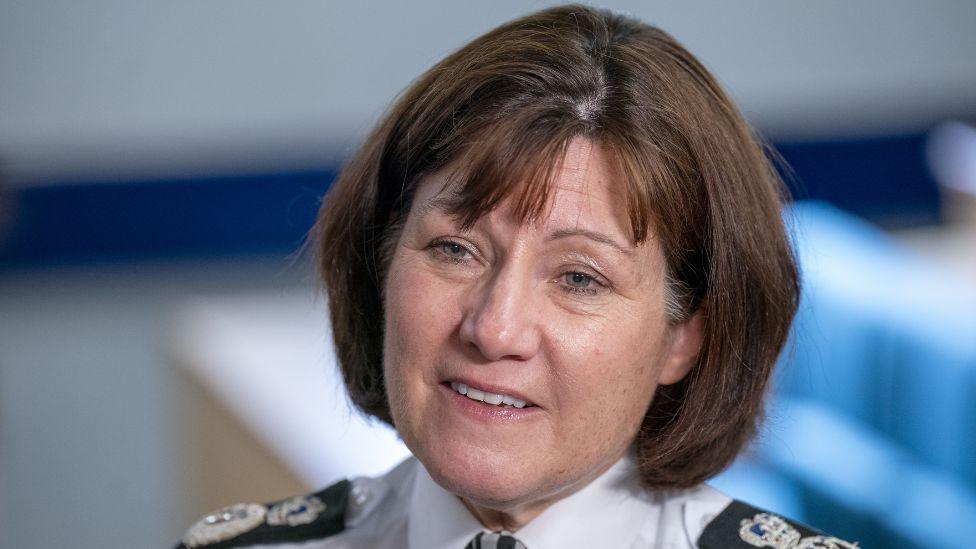 Joe Farrell, a woman with short brown hair, in a close-up shot. She is visible from the shoulders up and is wearing a white shirt with police badges on the shoulders