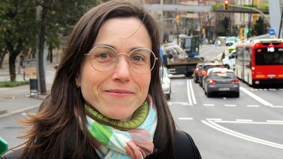 Maria, wearing glasses and a green polo necked jumper, stands in front of a busy road.