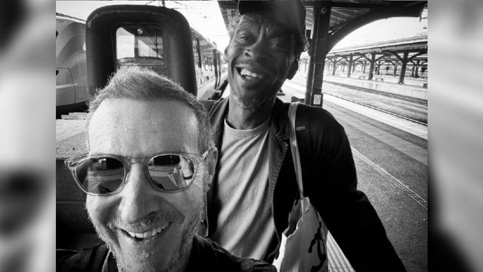 Massive Attack's two band members smiling in a black and white selfie at a train station. One is wearing sunglasses and the other a cap, while holding their luggage.