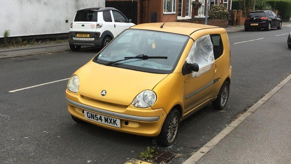 A bright yellow-gold Aixam car, parked slightly away from the kerb in a residential street. It is a two-door hatchback with a number plate from 2004.