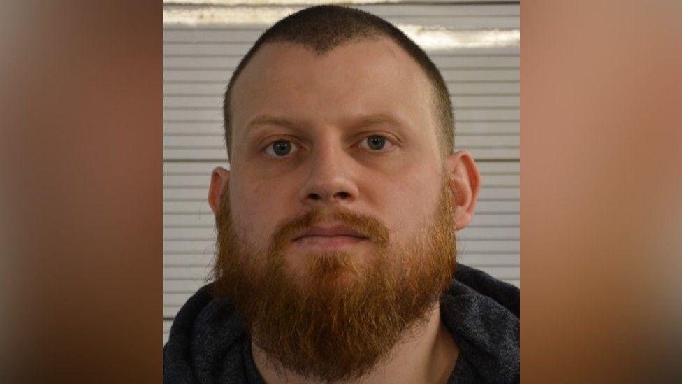 Mugshot of a man who has a long ginger beard, as well as a ginger moustache and short dark hair. He is staring into the camera and the hood of a grey hoodie can be seen. 