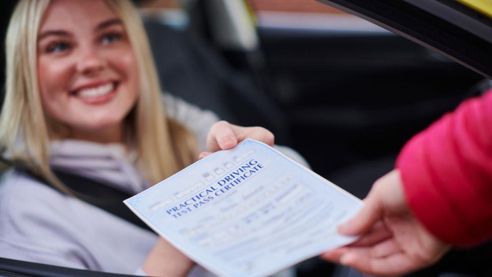 woman sitting in driving seat of car smiling as someone hands her a test pass certificate