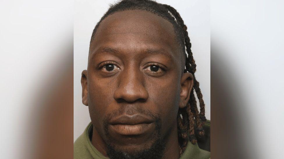 Shepherd Murombedzi looking at the camera in a police mugshot. He has long dreadlocks and is wearing a green top.