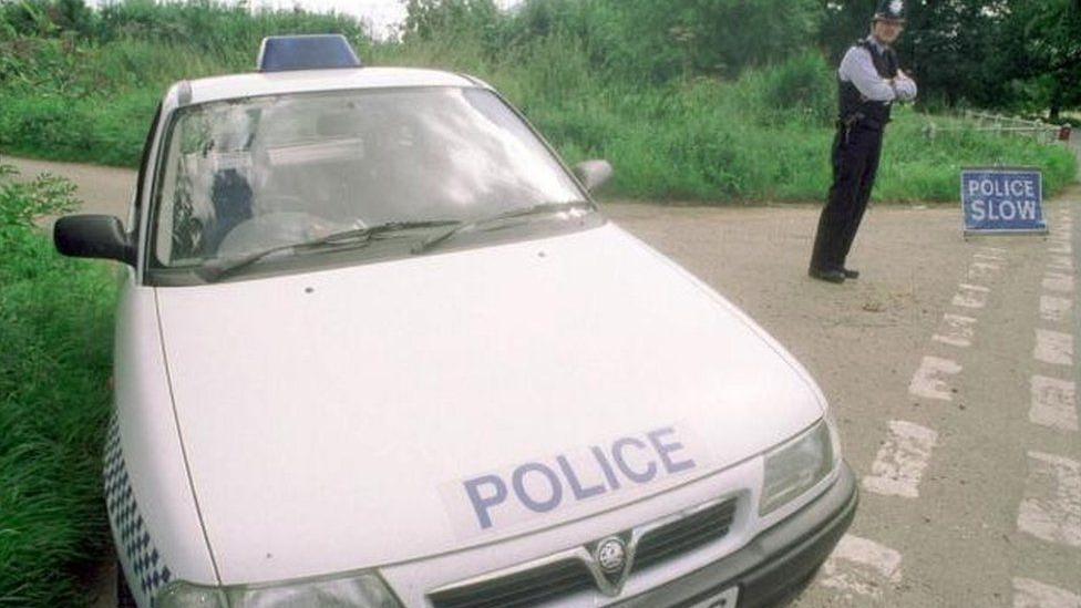 An old police car is parked at the top of a rural lane. It is white with the word "police" in blue on the bonnet. A police officer is standing nearby wearing a helmet. He has his arms crossed. There is also a sign on the road, warning drivers to go slowly near the scene