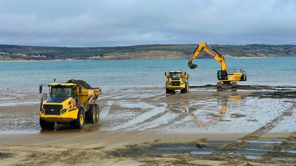 Yellow digger and two dumper trucks on sandy beach - the tide is out and pools of water have formed - cliffs and coastline can be seen across the sea