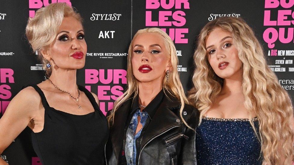 Orfeh, Christina Aguilera and Jess Folley standing next to each other on a red carpet in front of a Burlesque board. 
