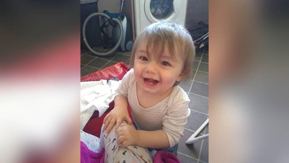 Young child smiles at the camera as he holds on to the person taking the photographs leg. There is a vacuum cleaner and a washing machine in the background of the image.