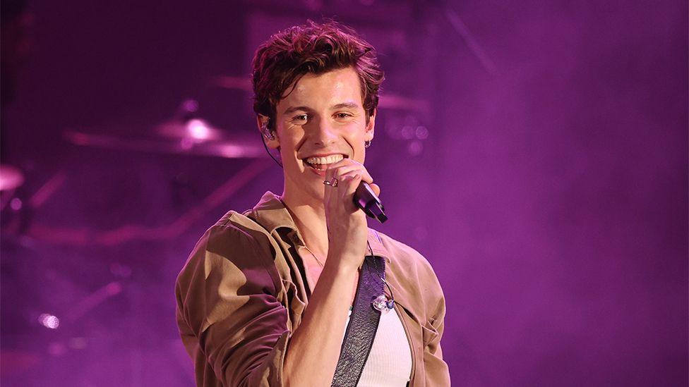 Shawn Mendes performing on stage, with a black microphone in his hand, a brown top with a white vest, and a purple haze background.