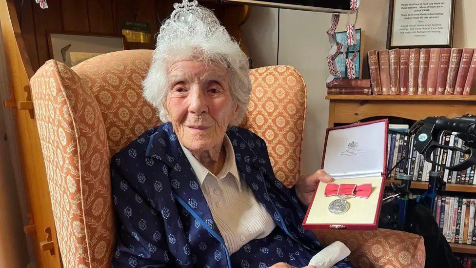 Joan Willett holds her BEM medal in a red box and sits in an armchair that is orange and patterned. Next to her chair there is a bookcase with books and DVDS on it. 