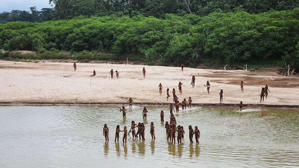 A group of people standing and wading in shallow water near the edge of a sandy riverbank. More individuals are scattered across the sandy area, with dense green forest in the background. The scene appears natural and remote, with no visible modern structures, and the water is calm and reflective.