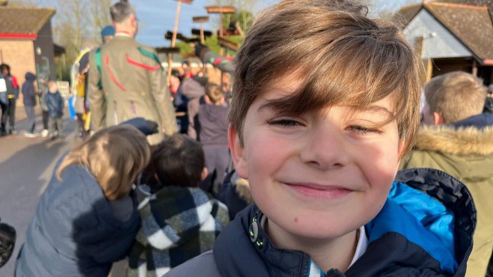 Koen is a young boy smiling at the camera. He has mid-brown hair sweeping in a fringe over his eyes. He is wearing a blue anorak and other children and dancers can be seen behind him in the playground