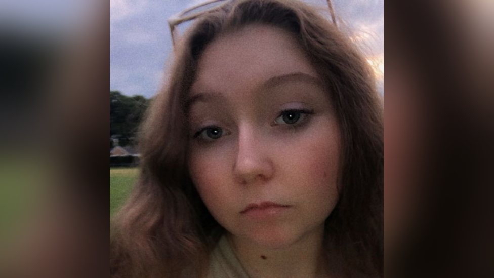 A selfie photograph of Erin with long dark hair and sunglasses on her head, looking directly into the camera with a serious expression.