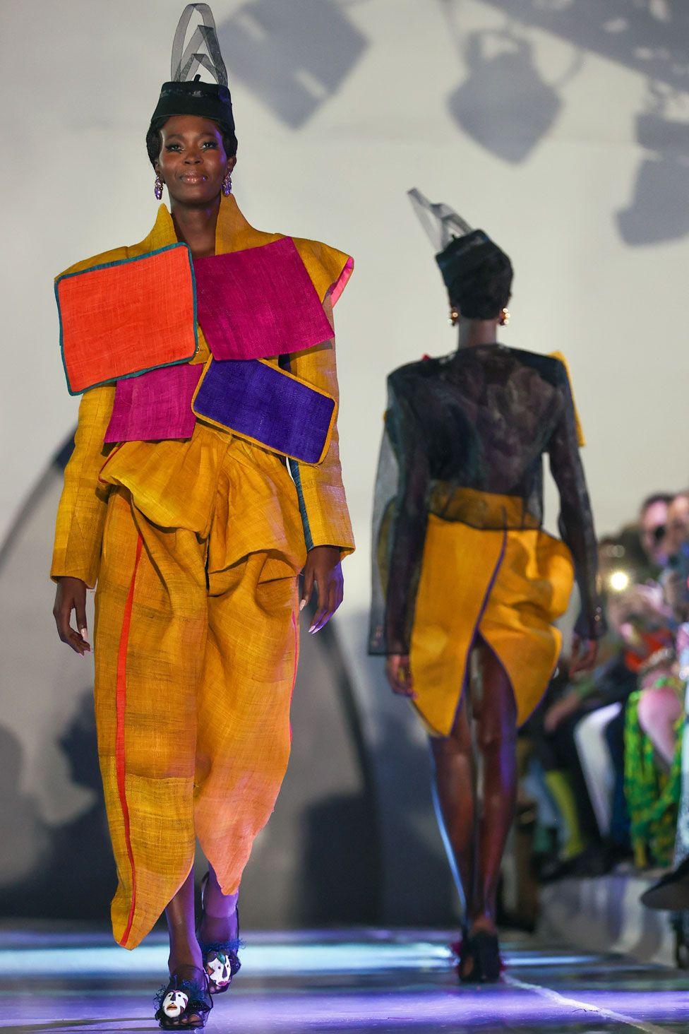 A model walks down the catwalk in a tailored linen two-piece suit. The colour is mustard and there are four rectangular panels stitched to the jacket, in magenta, orange and blue. In Dakar, Senegal - Saturday 7 December 2024